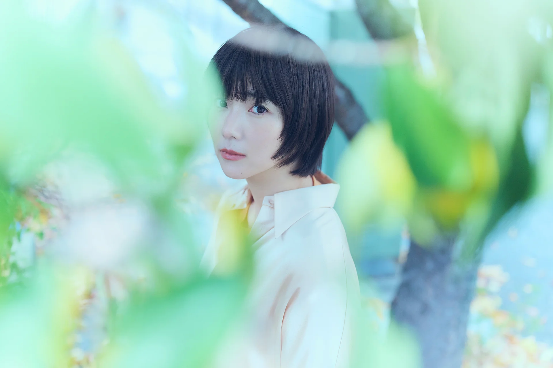 Oseba s kratkimi lasmi stoji na prostem, obkrožena z zelenimi listi.