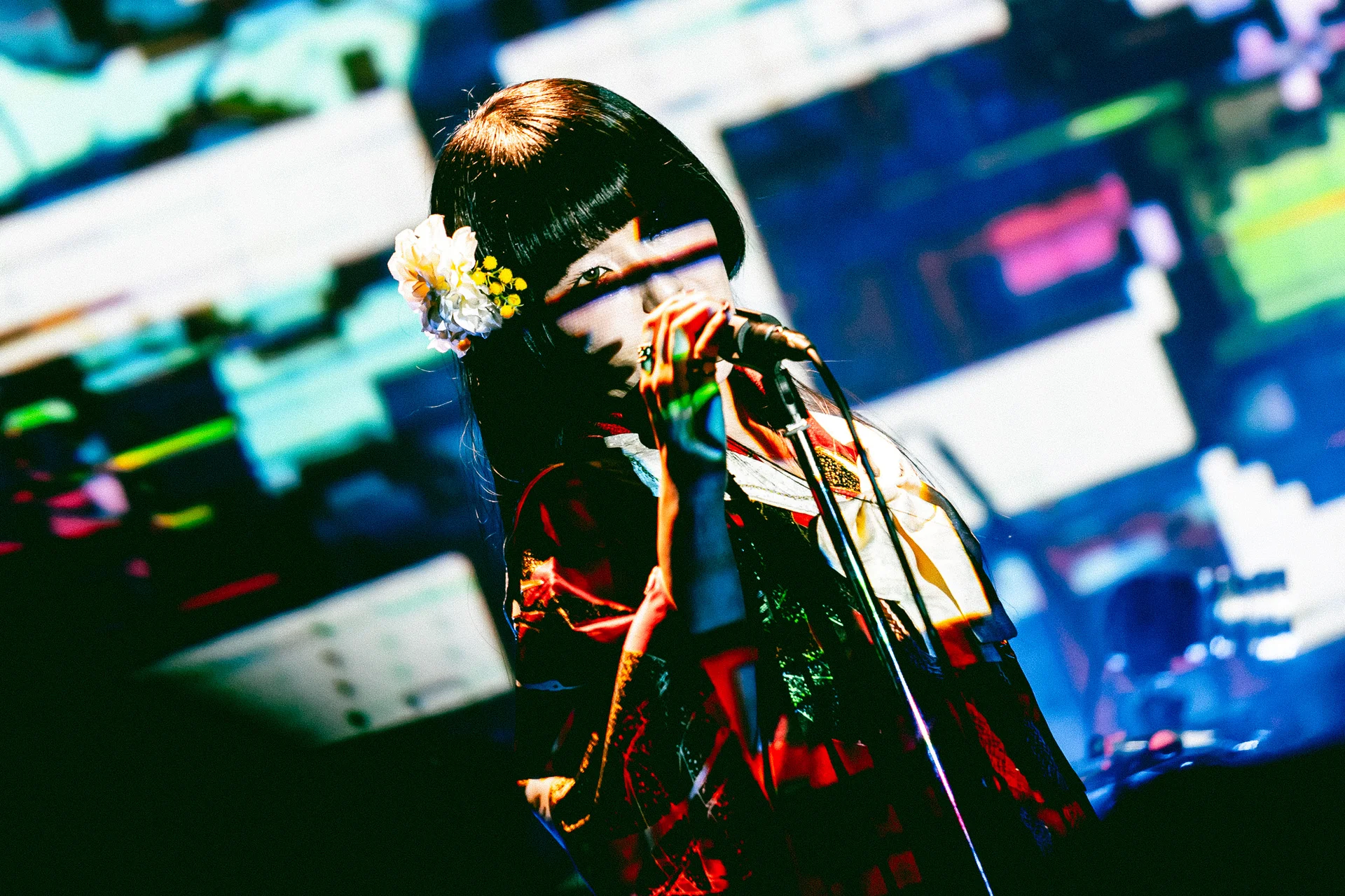 কিমোনো পরিহিত একজন শিল্পী মঞ্চে মাইক্রোফোন নিয়ে রঙিন ব্যাকগ্রাউন্ডে