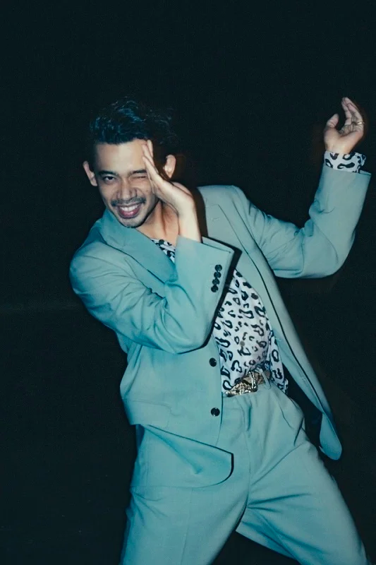 Man in a suit posing with a playful gesture against a dark background