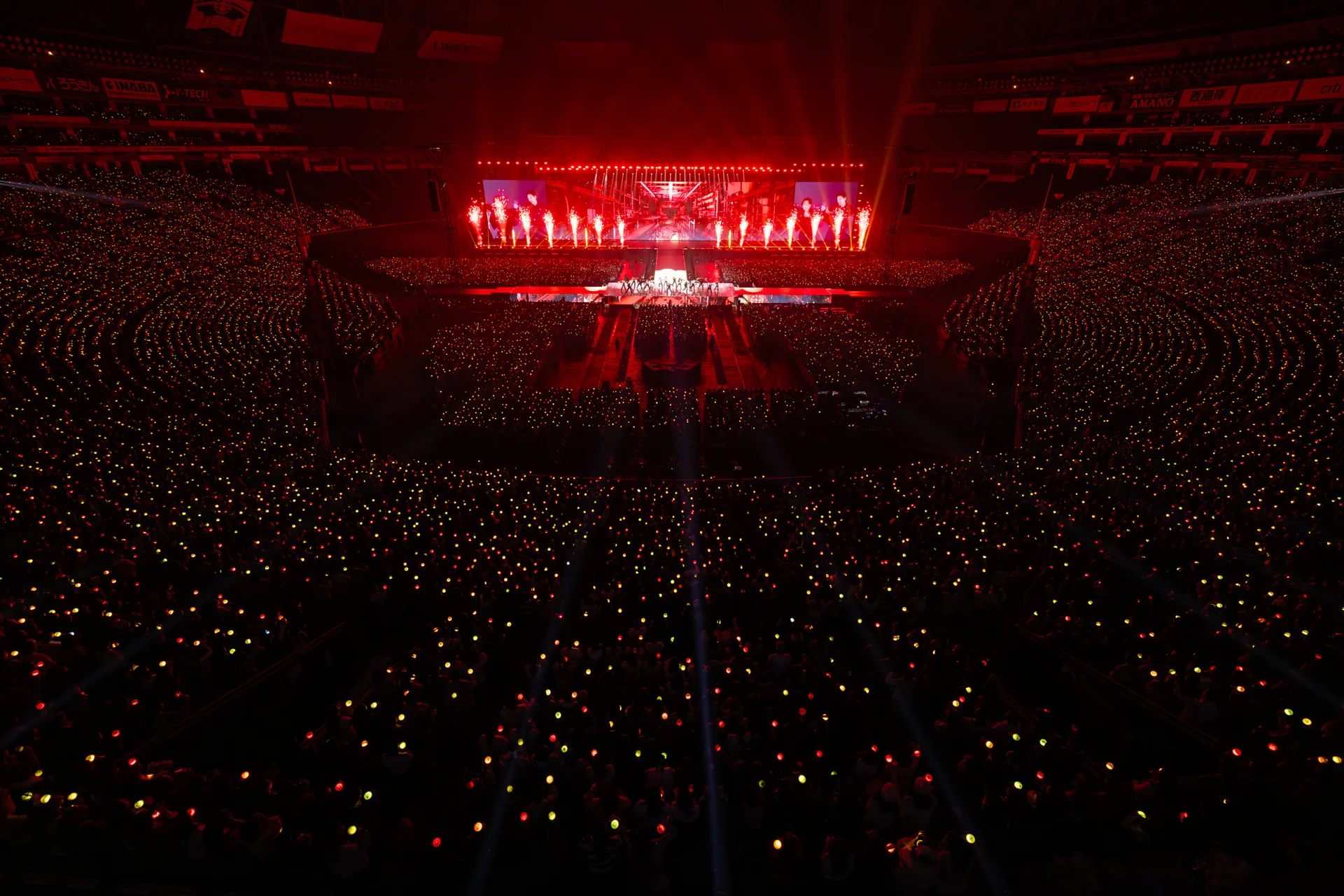 A concert stage with red lighting and a large audience waving light sticks.