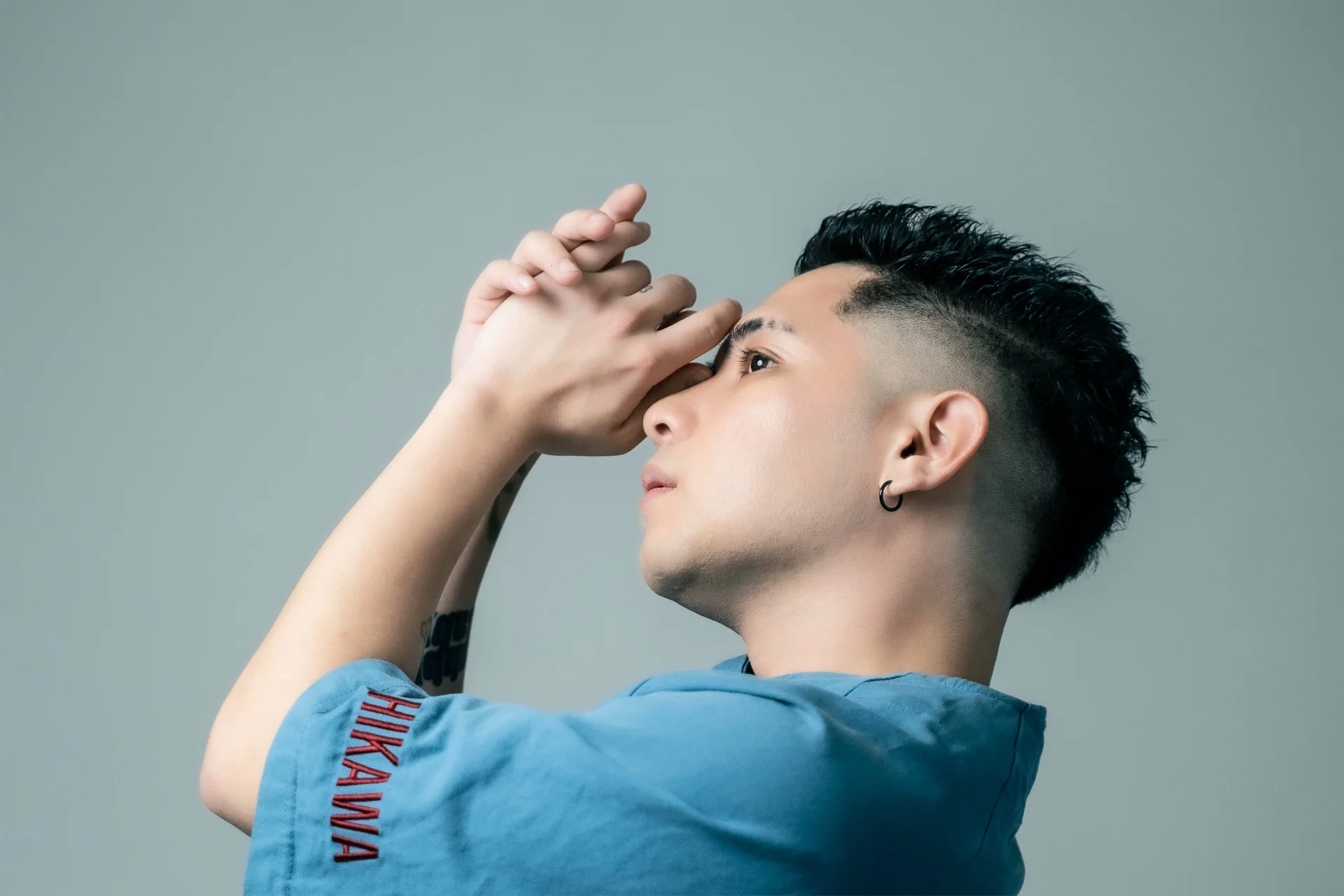 Person in blue shirt with HIKAWA text looking upwards with folded hands