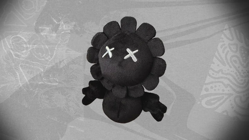 Plush toy with a black flower-like design and X-shaped eyes on a gray background