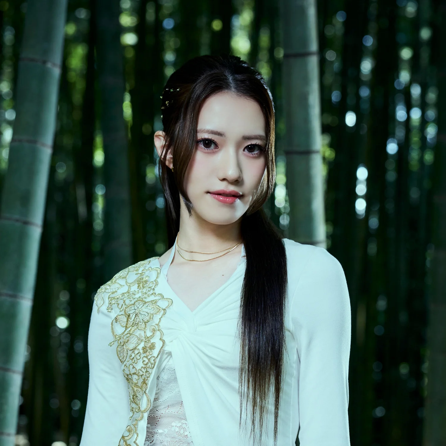 Млада жена со долга коса стои во бамбусова шума, носејќи бела облека со златни украси