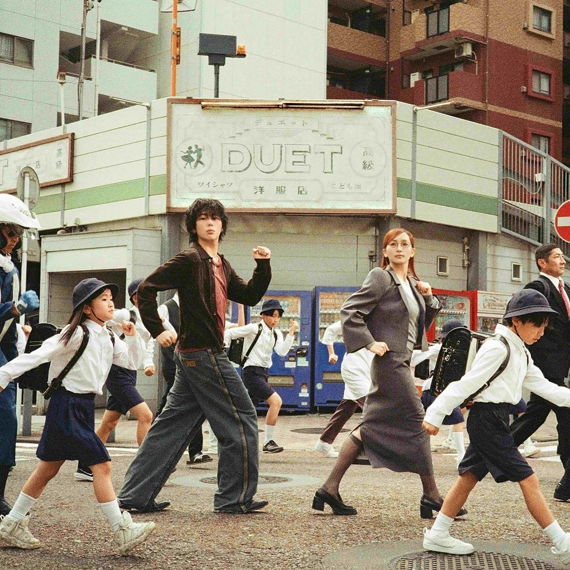 Ihmisiä, mukaan lukien univormuihin pukeutuneita lapsia, ylittämässä katua rakennuksen edessä, jossa on DUET-kyltti