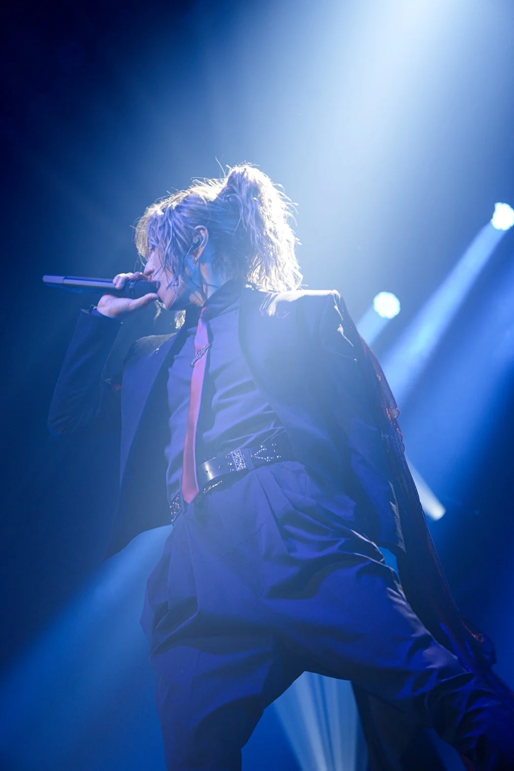 Artista cantando en el escenario bajo una iluminación dramática, vistiendo traje y corbata.