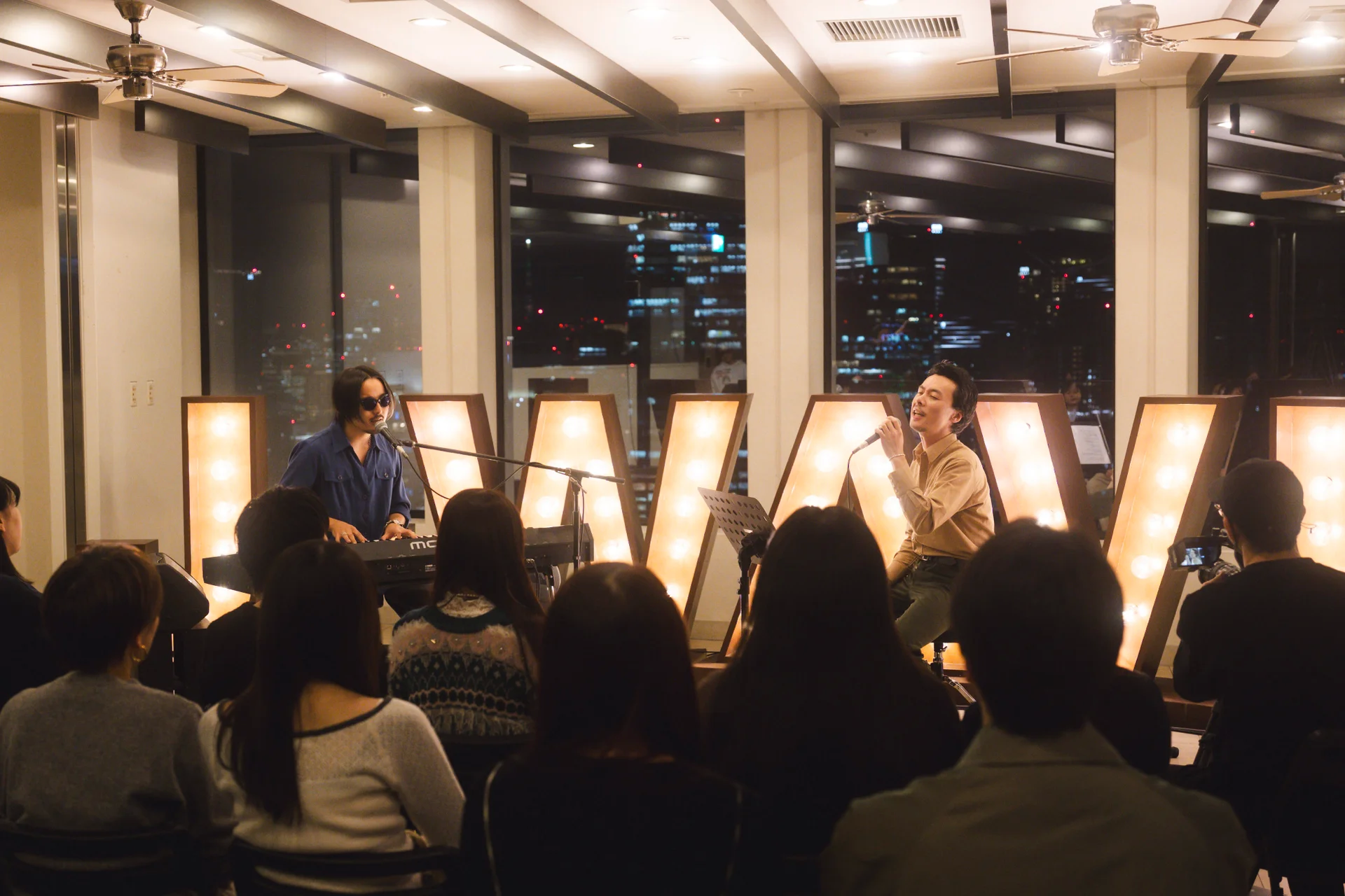 Perfformiad byw dan do gyda dau gerddor, cynulleidfa yn y blaen, arwydd J-WAVE o fwlbiau golau, cefndir dinas.