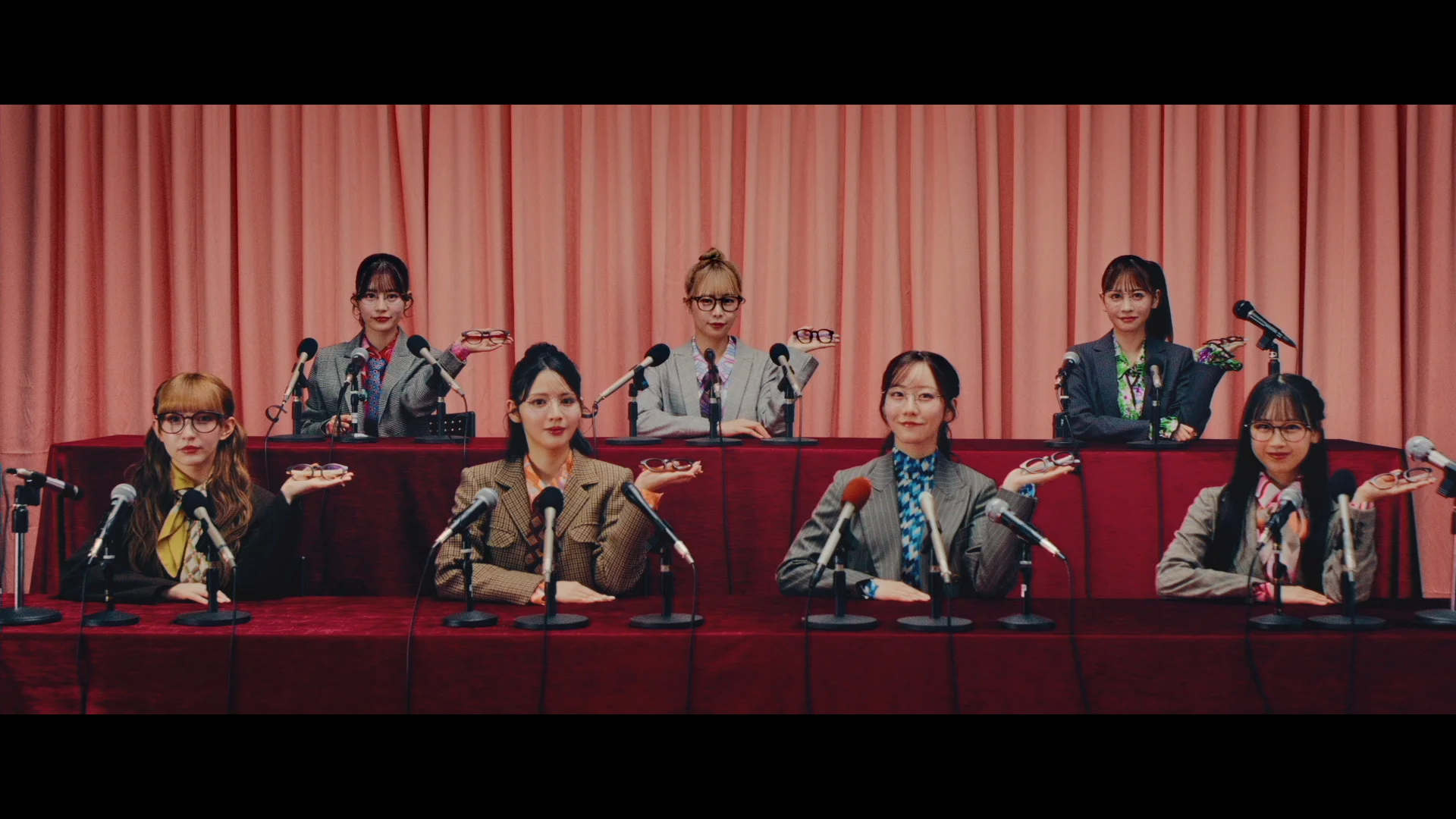 Grupa siedmiu kobiet w garniturach siedzi przy stole z mikrofonami, pozując z figlarnymi wyrazami twarzy i gestami