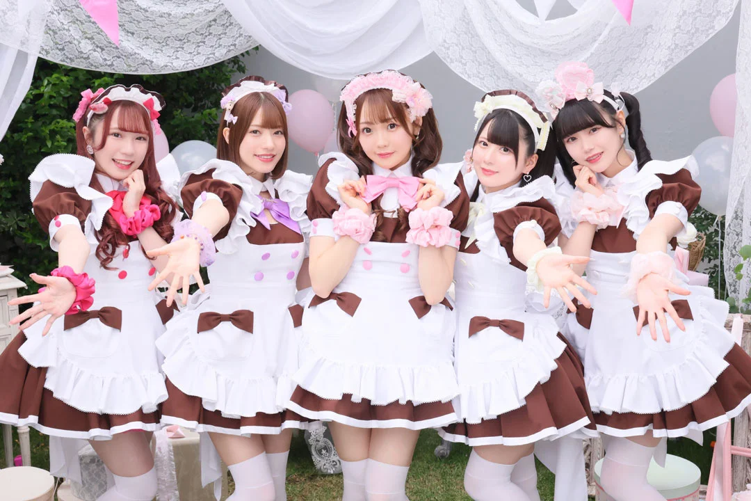 Fem kvinner i maid-drakter fra Kanzen Maid Sengen smiler med åpne armer