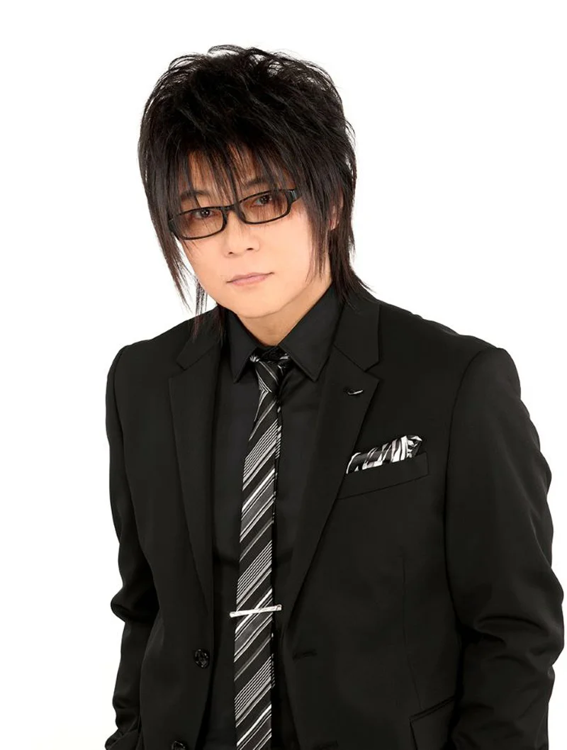 Il doppiatore Morikawa Toshiyuki in completo nero con occhiali su sfondo bianco