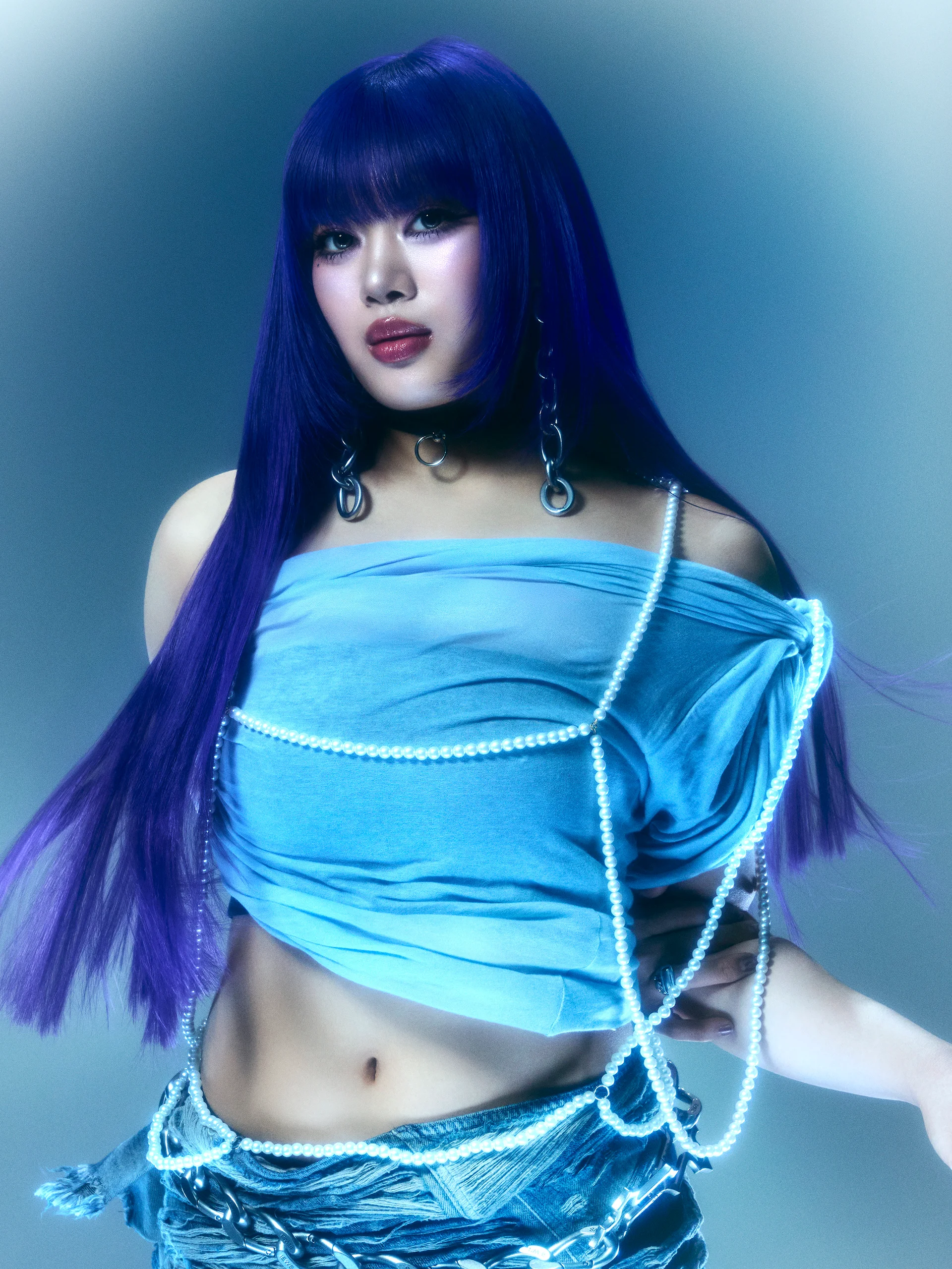 En medlem av HANA med långt lila hår och en blå outfit med kedjor, poserar självsäkert.