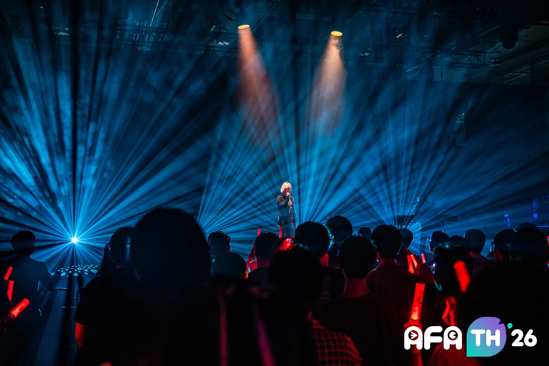 ReoNa performing on stage at AFA Thailand 2026 under blue spotlights, audience holding red glow sticks.