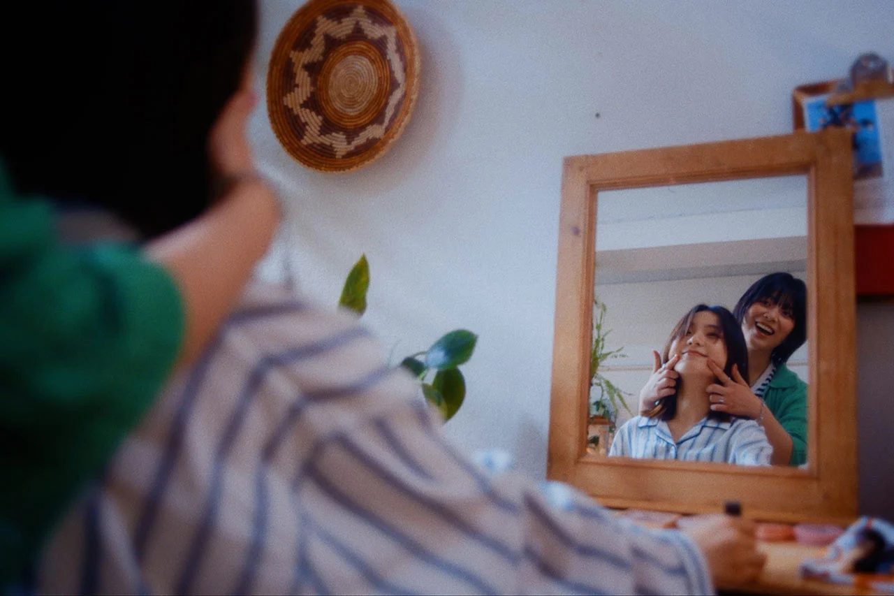 Two people smiling in front of a mirror, one playfully pulling the other