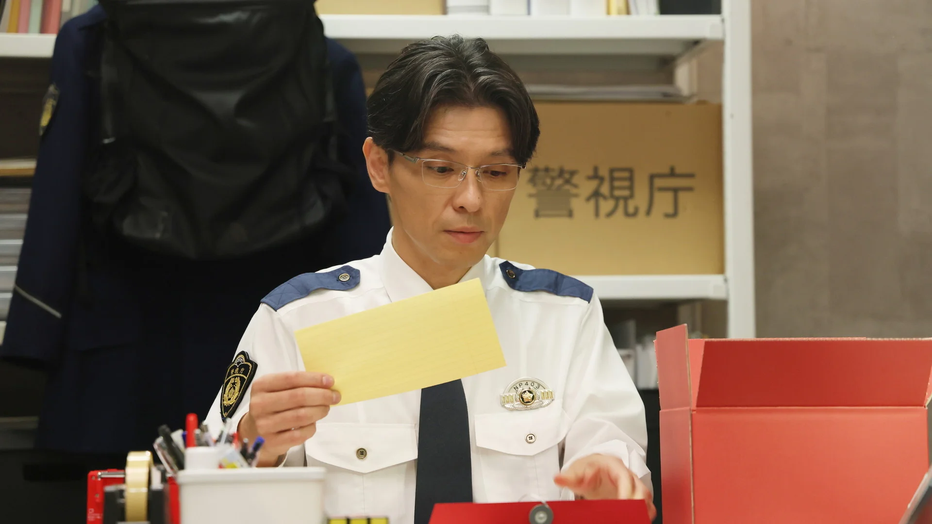 Un home amb uniforme de policia seua a un escriptori examinant una carpeta, envoltat de material de oficina.