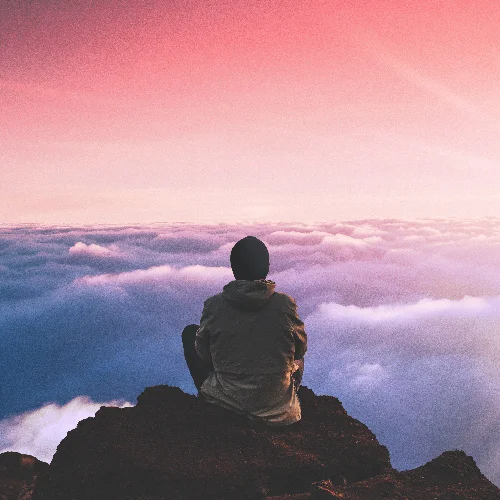 Person sitting on a cliff overlooking clouds at sunset